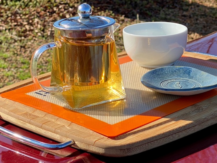 picture of yellow hued tea in a clear tea pot with an infuser and a blue saucer and a white bowl all set upon a wooden serving tray on top of a red cooler outside