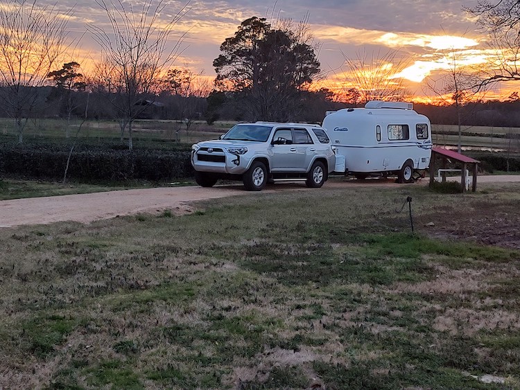 picture of a silver suv car pulling a small white camper on a dirt road with a sunset in the background